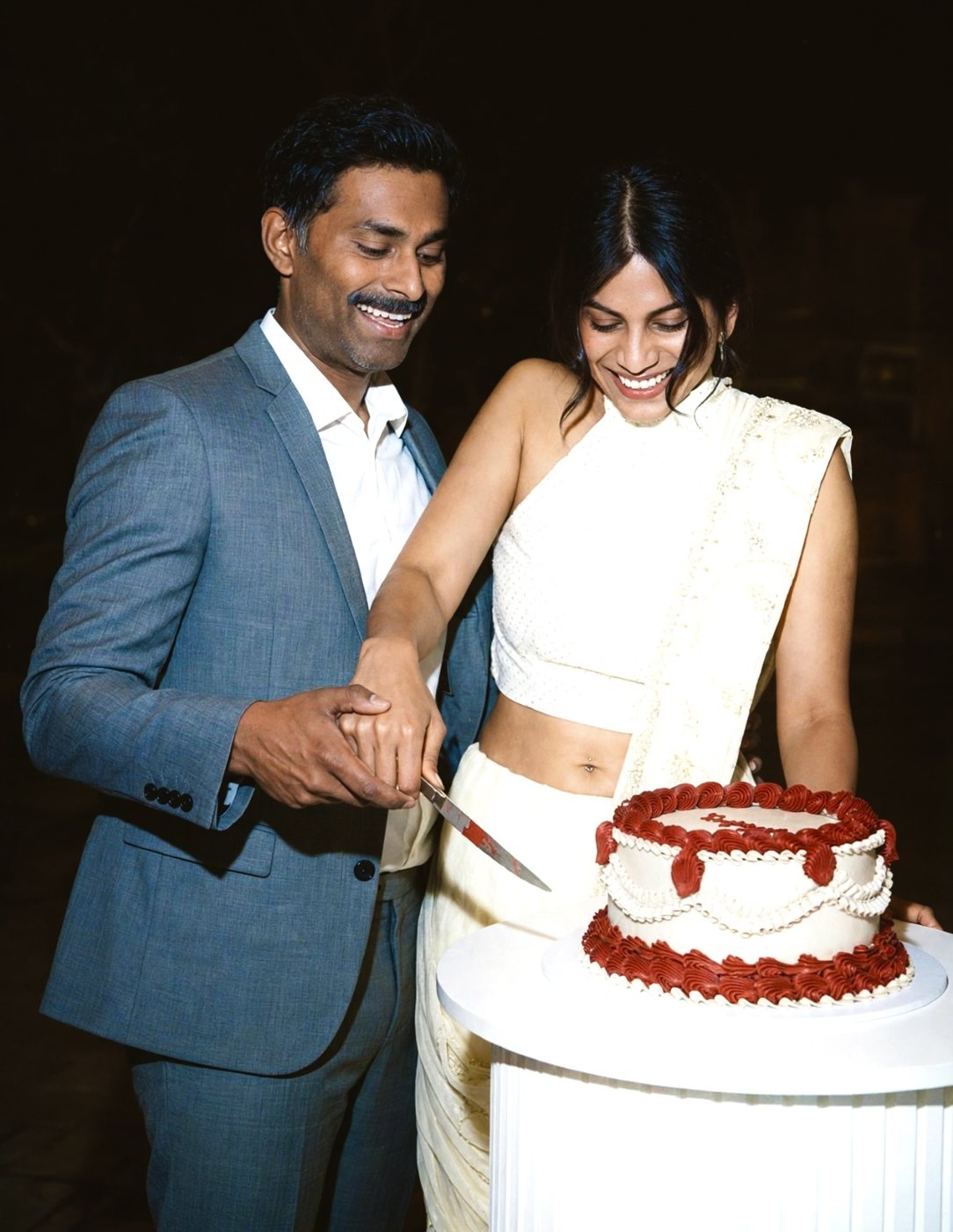 Randi and Sudesh smiling as they cut a red and white cake together
